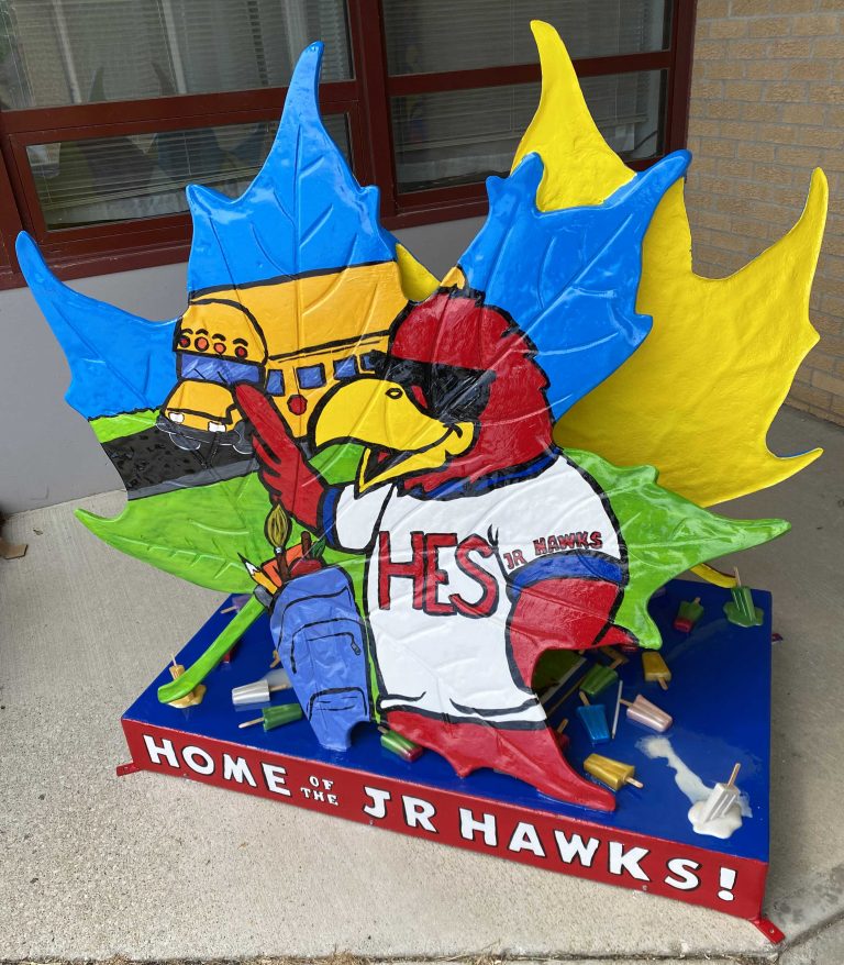A fiber glass maple leaf on a small platform featuring the Hiawatha Red Hawk mascot wearing sunglasses, with a school bus behind the Red Hawk.