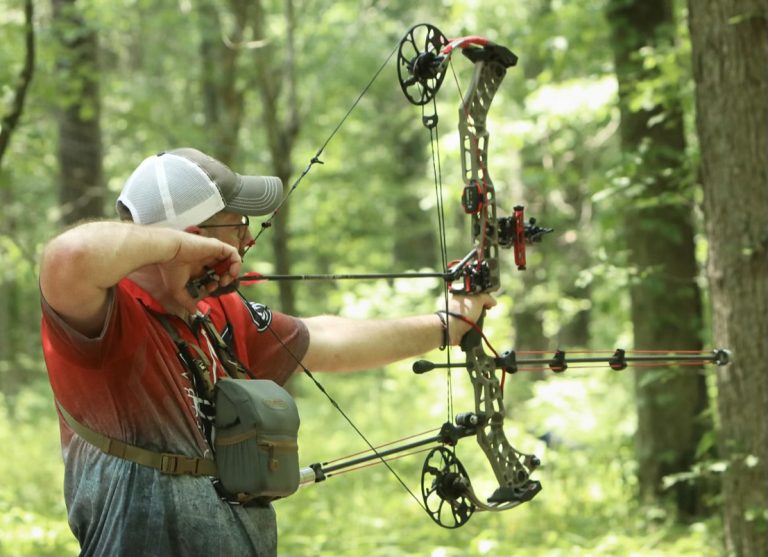 A man with a bow taking aim to let loose an arrow in a green forest backdrop.