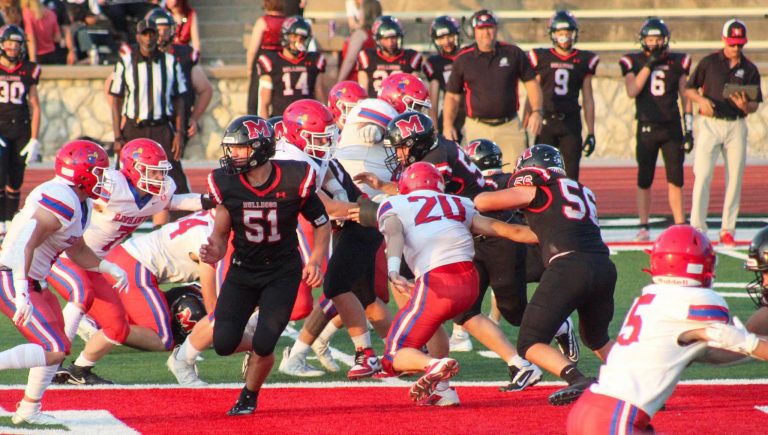 Two teams of football--one in red and white uniforms and the other in black uniforms--tackle each other on a football field in front of a crowd.