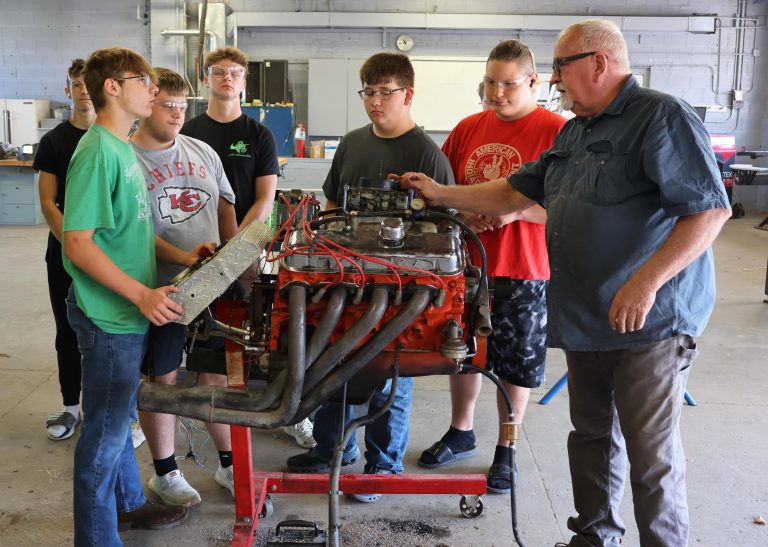 A group of people--including a teacher--around some mechanical parts in a workshop setting.