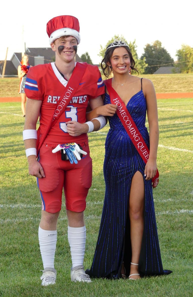 A high school boy in a red uniform wears a red crown and a high school girl in a blue dress with a red sash and a crown stand in a green football field.