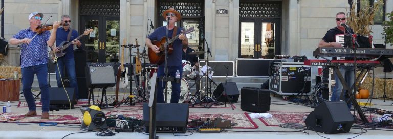 A country rock band in front of a courthouse with stereo equipment present.