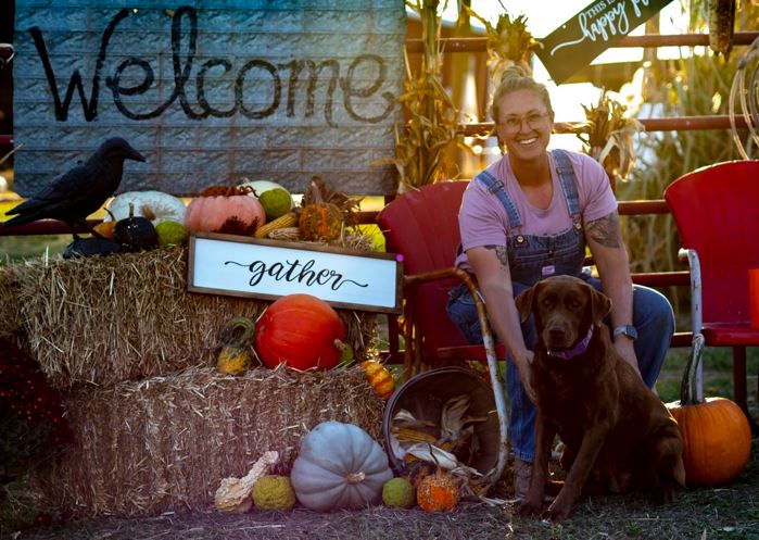 A woman and a dog pose for a photo in a farm setting with pumpkins and haystacks.