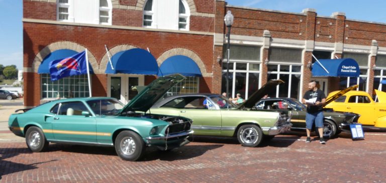 A row of cars in daylight in front of some buildings. The vehicles are on a cobblestone street.