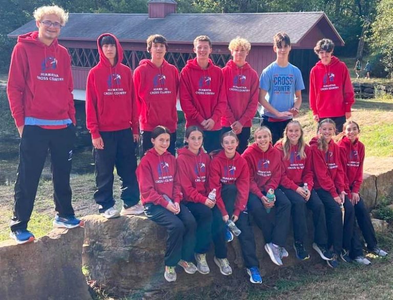 A group of young cross country runners, all but one in red hoodies with the Red Hawk logo standing in front of a building; the back row of boys is standing while the bottom row of girls is sitting.