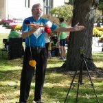 A gray haired man holds up a contraption with balls on a string attached to a pole on a sunny day; there is also a tripod with a smartphone attached to his right.