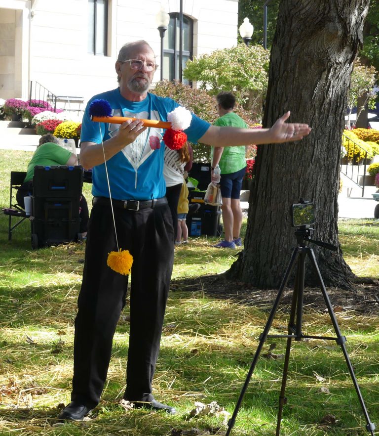A gray haired man holds up a contraption with balls on a string attached to a pole on a sunny day; there is also a tripod with a smartphone attached to his right.