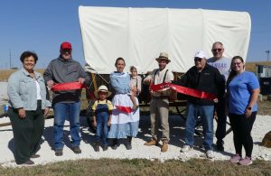 A group of people in front of an old wagon with a white cover during the day.