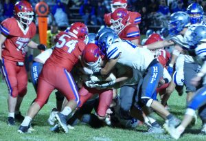 A group of football players--one in red and the other in white and blue--tackle each other on a green football field at night.